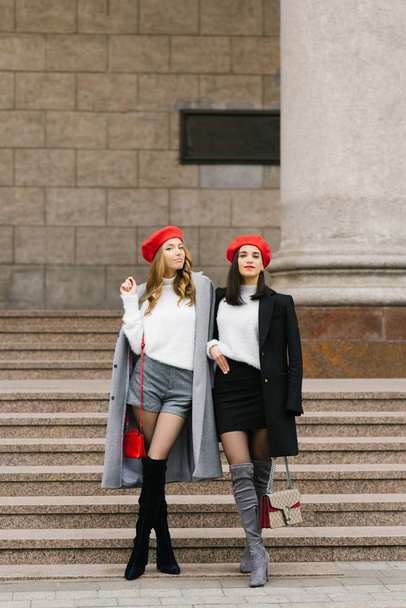 Two beautiful French women in berets, looking at the camera and smiling while walking together in the open air near the steps of a classic building. - Photo, Image