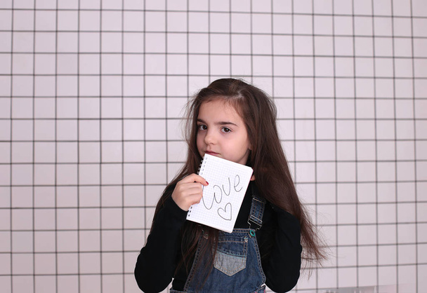 jong lachend meisje van acht jaar met lang haar in een zwarte coltrui en denim sundress met een teken "liefde en hart" - Foto, afbeelding