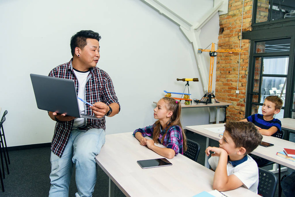 Ein asiatischer Lehrer sitzt mit Laptop in der Hand am Schreibtisch und erklärt sechs Grundschülern den Unterricht. Schüler sitzen am Schreibtisch und hören ihrem Dozenten zu. - Foto, Bild