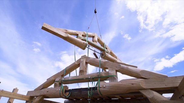 Kran über dem im Bau befindlichen Holzgebäude an einem sonnigen Tag im Grünen. Clip. Holzbalkenkonstruktion wird über den Hausrahmen transportiert. - Foto, Bild
