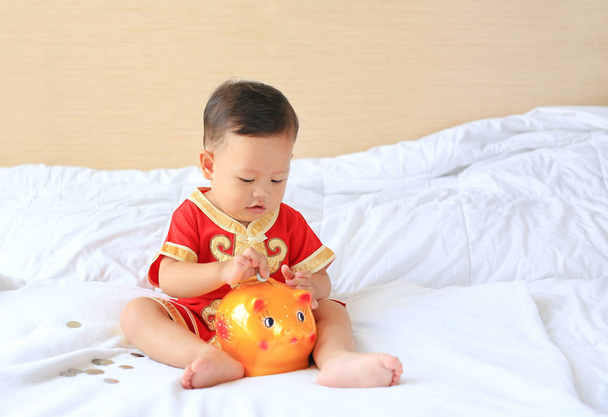 Little Asian baby boy in traditional Chinese dress putting some coins into a piggy bank sitting on bed at home. Kid saving money concept. - Photo, image