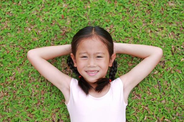 Sonriente niña asiática con dos cabellos de cola de caballo acostado sobre hierba verde en el jardín
. - Foto, imagen