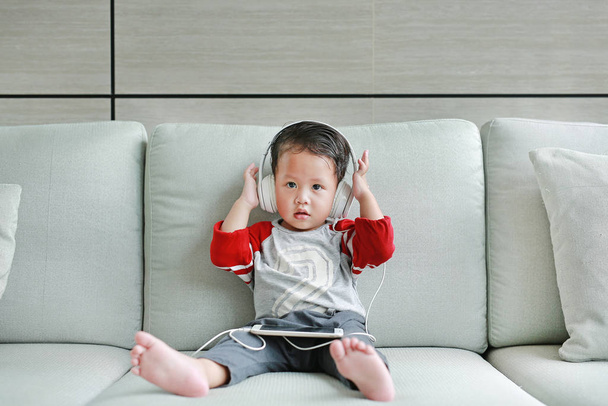 Adorable little Asian baby boy in headphones is using a smartphone lying on the sofa at home. Child listening to music on earphones. - Fotó, kép