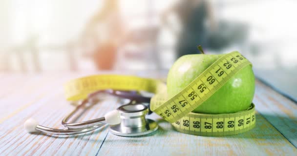 zdrowy tryb życia i odżywianie - jabłko z taśmą pomiarową i stetoskopem na stole w siłowni z biegającymi ludźmi w tle - Materiał filmowy, wideo