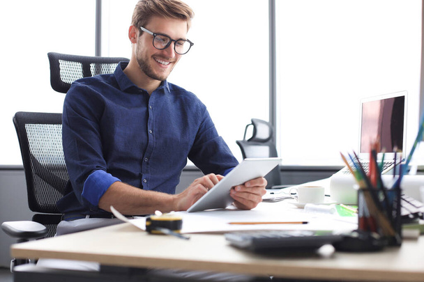 Νεαρός σύγχρονος επιχειρηματίας που εργάζεται χρησιμοποιώντας ψηφιακή ταμπλέτα ενώ κάθεται στο γραφείο. - Φωτογραφία, εικόνα