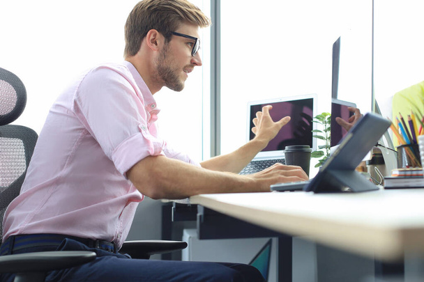 Homem de negócios jovem pensativo na camisa que trabalha usando o computador ao sentar-se no escritório
. - Foto, Imagem