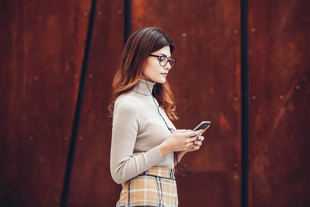 Image of beautiful stylish woman holding mobile phone. Young european girl standing at the street and using cellphone. Woman listening to music with the phone and having fun. Mobile internet concept. Social media. - Photo, image