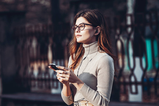 Εικόνα της όμορφης κομψής γυναίκας που κρατά το κινητό τηλέφωνο. Νεαρή Ευρωπαία στέκεται στο δρόμο και χρησιμοποιεί κινητό. Γυναίκα ακούει μουσική με το τηλέφωνο και διασκεδάζει. Κινητό διαδίκτυο έννοια. Μέσα κοινωνικής δικτύωσης. - Φωτογραφία, εικόνα