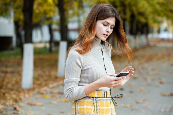 Kuva kaunis tyylikäs nainen tilalla matkapuhelin. Nuori eurooppalainen tyttö seisoo kadulla ja käyttää kännykkää. Nainen kuuntelee musiikkia puhelimen kanssa ja pitää hauskaa. Mobiili internet käsite. Sosiaalinen media
. - Valokuva, kuva