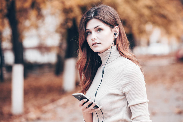 Image of beautiful stylish woman holding mobile phone. Young european girl standing at the street and using cellphone. Woman listening to music with the phone and having fun. Mobile internet concept. Social media. - Photo, Image