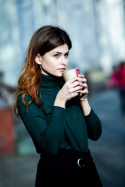 Young stylish woman drinking tea in a city street. Hipster european girl with latte paper glass  in street. Gorgeous young woman with cup of coffee in city street. Coffee break. Coffee to go.  - 写真・画像
