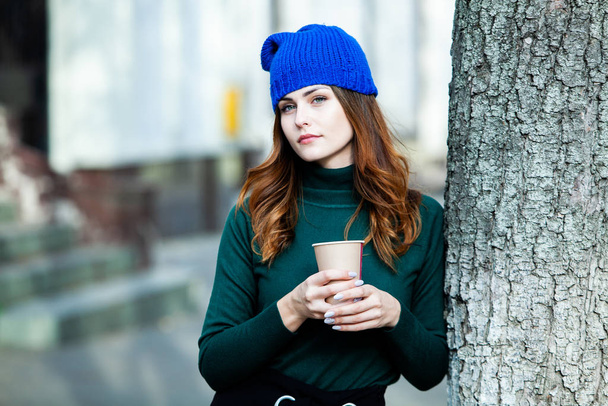 Young stylish woman drinking tea in a city street. Hipster european girl with latte paper glass in street. Gorgeous young woman with cup of coffee in city street. Coffee break. Coffee to go.  - Photo, Image