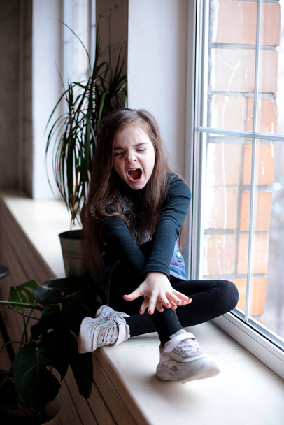 young smiling girl of eight years with long hair in a black turtleneck and jeans sundress walks on the windowsill near the window in a good mood different emotions - Photo, Image
