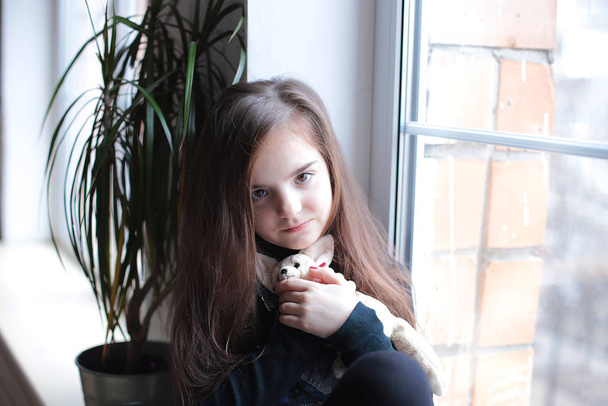 νεαρό χαμογελαστό κορίτσι οκτώ ετών με μακριά μαλλιά σε μαύρο ζιβάγκο και τζιν sundress βόλτες στο περβάζι του παραθύρου κοντά στο παράθυρο σε καλή διάθεση διαφορετικά συναισθήματα - Φωτογραφία, εικόνα