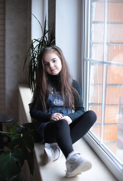 young smiling girl of eight years with long hair in a black turtleneck and jeans sundress walks on the windowsill near the window in a good mood different emotions - 写真・画像