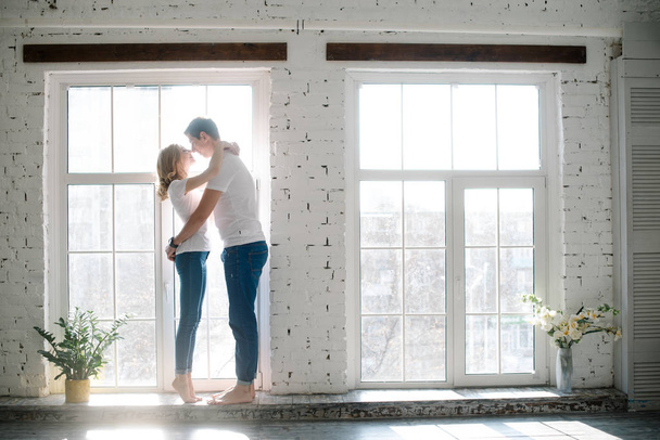 petits câlins d'un jeune couple marié serrant et regardant dans les yeux de l'autre et touchant le nez près d'une grande fenêtre à la lumière du matin à la maison - Photo, image