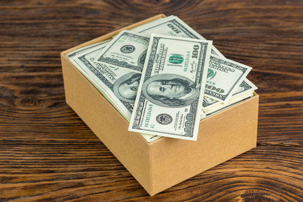 μικρό χαρτονένιο κουτί γεμάτο τραπεζογραμμάτια των εκατό δολαρίων close-up με επιλεκτική εστίαση σε φυσικό φόντο ξύλο - Φωτογραφία, εικόνα
