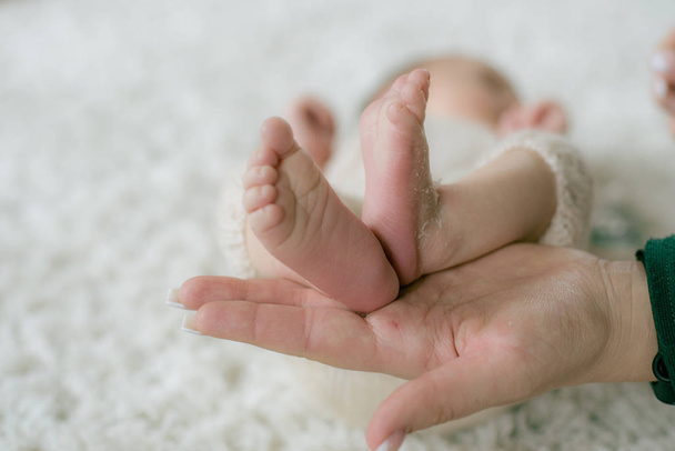 Annenin elleri, evde örgü ekose kumaşlarla hasır bir sepetin içinde yatan bir bebeğin küçük sevimli bacaklarını tutuyor. Mutlu annelik - Fotoğraf, Görsel