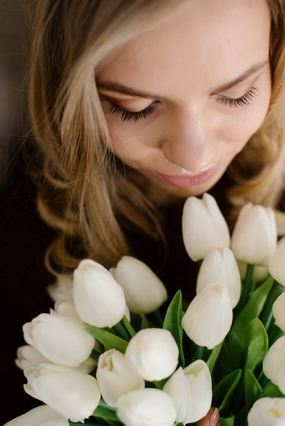 portrait of a young blonde girl looking down smelling flowers white tulips bouquet close-up view from above in a photo studio, thick long eyelashes and gentle light makeup are visible - Zdjęcie, obraz