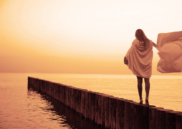 evening or Morning walk of pretty blond woman. Caucasian blonde girl walking on sunset beach. log direct way to horizon . rear, back view. white wet towel on shoulders  - Φωτογραφία, εικόνα