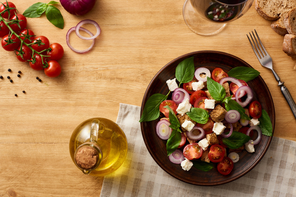 vrchní pohled na vynikající italský zeleninový salát panzanella podávané na talíři na dřevěném stole v blízkosti čerstvých surovin a červené víno - Fotografie, Obrázek