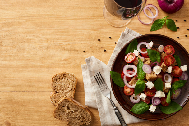 top view of delicious Italian vegetable salad panzanella served on plate on wooden table near fresh ingredients, fork, bread and red wine - 写真・画像
