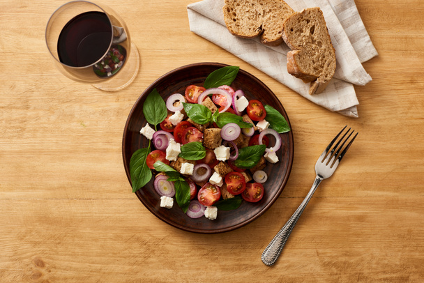 top view of delicious Italian vegetable salad panzanella served on plate on wooden table near fresh bread and red wine - Photo, Image