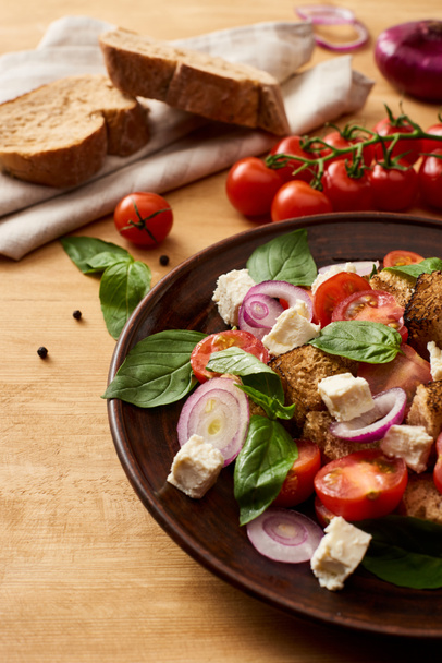 delicious Italian vegetable salad panzanella served on plate on wooden table near fresh ingredients and bread - Photo, Image