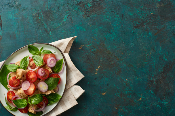 сверху вид на вкусный итальянский овощной салат panzanella подается на тарелке на текстурированной зеленой поверхности с салфеткой
 - Фото, изображение