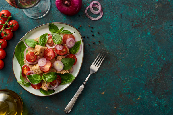 top view of delicious Italian vegetable salad panzanella served on plate on textured green surface with ingredients and fork - Foto, afbeelding