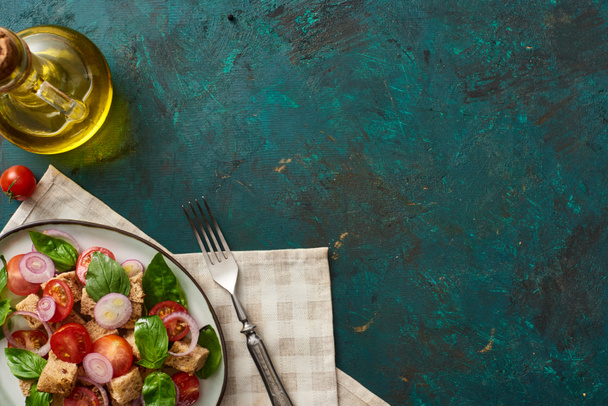 vista superior de la deliciosa ensalada de verduras italiana panzanella servido en el plato en la superficie verde con textura con aceite de oliva, servilleta y tenedor
 - Foto, Imagen