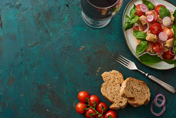 top view of delicious Italian vegetable salad panzanella served on plate on textured green surface with tomatoes, bread, red wine and fork - 写真・画像