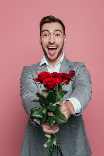 handsome excited man yelling and holding red roses, isolated on pink - Φωτογραφία, εικόνα