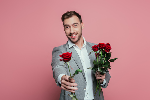 przystojny pozytywny mężczyzna w garnitur gospodarstwa czerwone róże kwiaty odizolowany na różowy - Zdjęcie, obraz