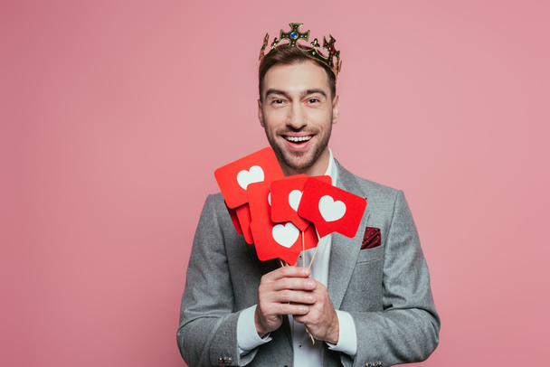 veselý muž v koruně drží karty se srdcem na Valentýna, izolované na růžové  - Fotografie, Obrázek