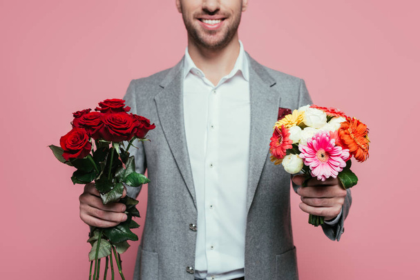 ostříhaný pohled na šťastného muže v obleku držícího dvě kytice, izolovaný na růžové - Fotografie, Obrázek
