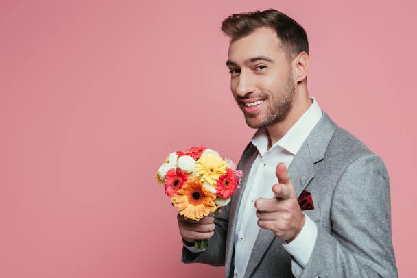 vousatý šťastný muž v obleku drží květiny a ukazuje na vás, izolovaný na růžové - Fotografie, Obrázek