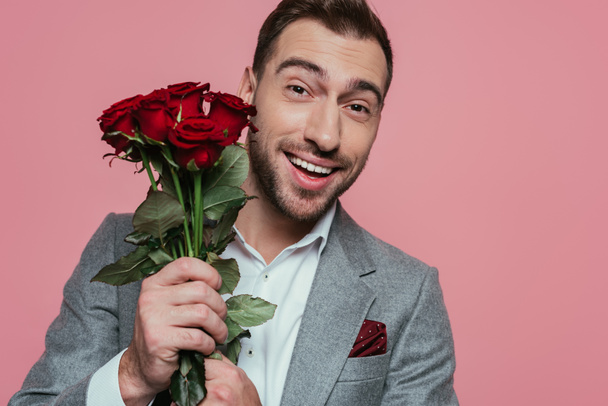 excited handsome man in suit holding roses, isolated on pink - 写真・画像