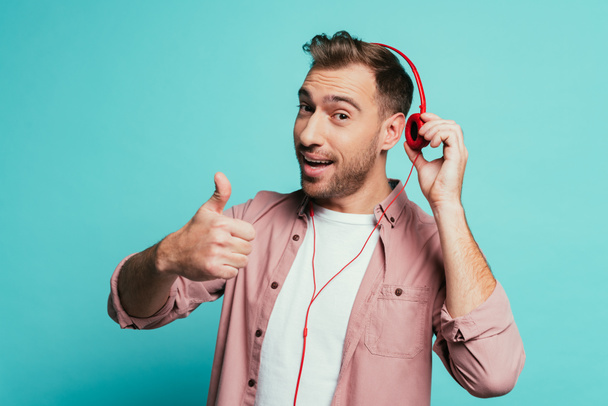 happy man listening music with headphones and showing thumb up, isolated on blue - Foto, afbeelding