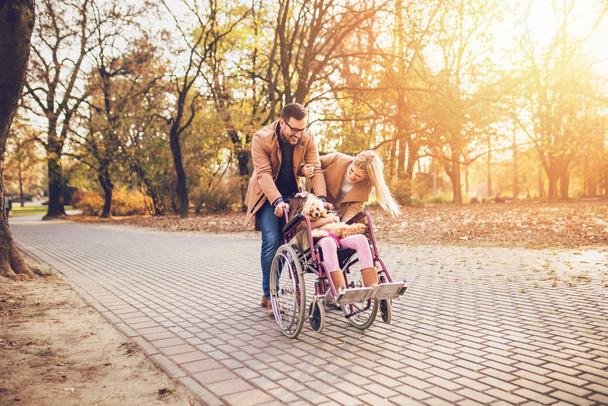 Девочка-инвалид в инвалидной коляске наслаждается прогулками с мамой, папой и собакой в парке
. - Фото, изображение