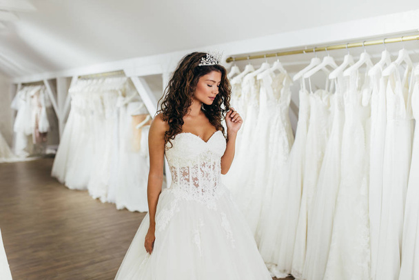Beautiful young brunette woman choosing wedding dress in a bridal salon. - Фото, зображення