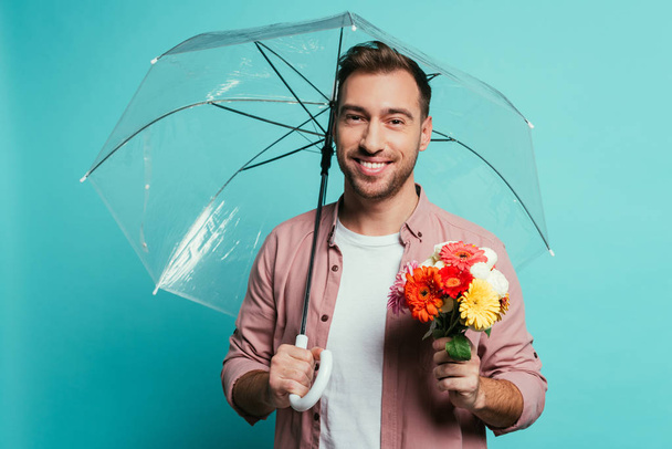 bärtiger lächelnder Mann mit Blumenstrauß und Regenschirm, isoliert auf blauem Grund - Foto, Bild