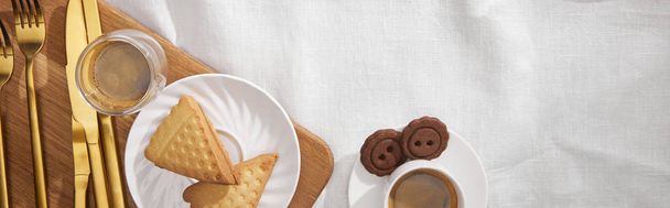 Bovenaanzicht van koffie met koekjes en bestek op snijplank op witte doek, panoramisch schot - Foto, afbeelding
