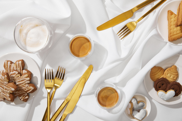 Bovenaanzicht van verse koekjes met glazuur, glazen koffie en bestek op golvend wit tafelkleed - Foto, afbeelding