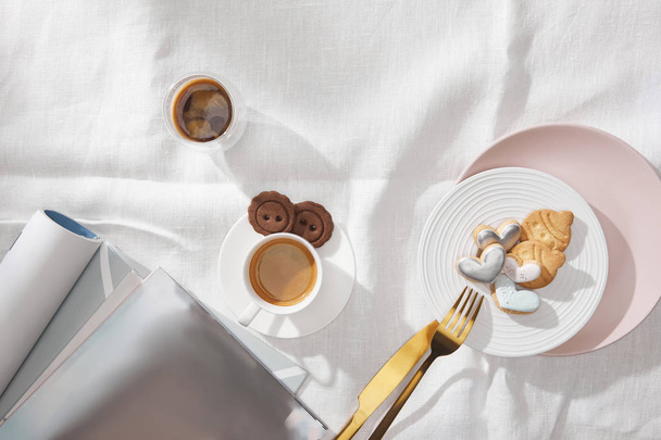 Draufsicht auf frischen Kaffee, Kekse mit Glasur und Zeitschriften auf weißer Tischdecke - Foto, Bild