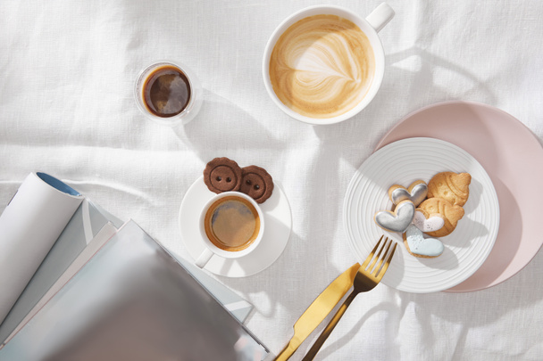 白いテーブルクロスに雑誌の横に新鮮なコーヒーとおいしいクッキーのトップビュー - 写真・画像
