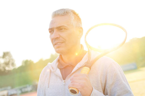 Mature athlete playing tennis in court with yellow lens flare in background  - Foto, Imagen