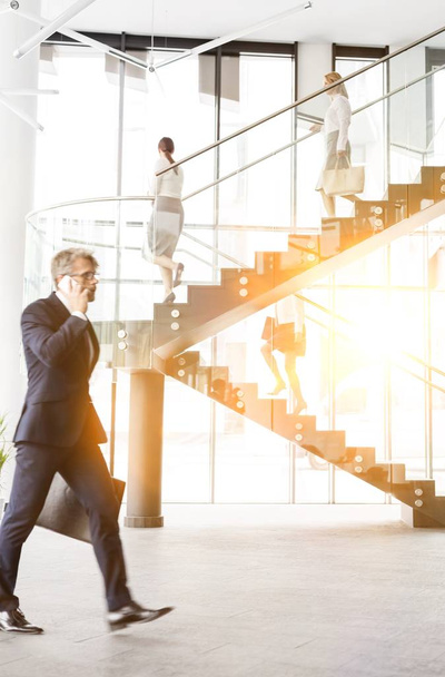 Mature businessman walking while talking on smartphone in office with yellow lens flare  - Photo, Image