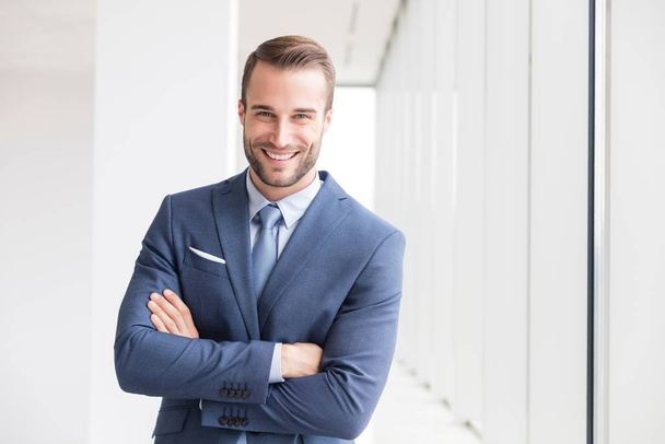 Portrait of smiling handsome young businessman standing with arms crossed in new office - 写真・画像