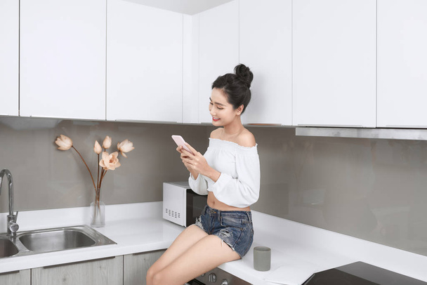Young beautiful Asian woman using smartphone and smiling while sitting in kitchen - Photo, image